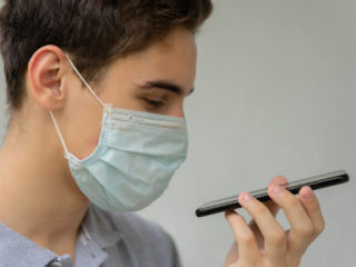 A young man in a disposable medical protective mask on his face is talking on the phone. Teen hold phone speak on speakerphone using virtual digital voice recognition assistant search.